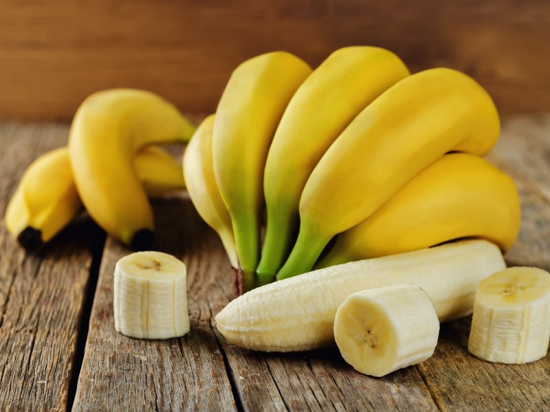 Sind Bananen gesund: Bananen mit und ohne Schale, einige geschnitten, auf Holz.