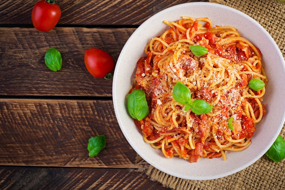 Ein Teller Spaghetti alla Amatriciana mit frischen Basilikumblättern in der Draufsicht. Daneben liegen Tomaten und Basilikumblätter.