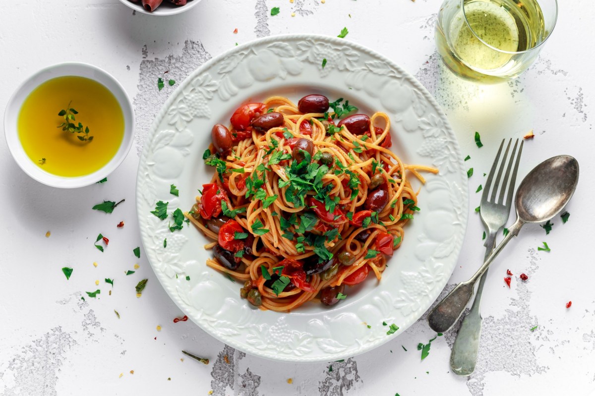 Spaghetti alla Puttanesca auf einem weißen, gemusterten Teller. Garniert sind sie mit Petersilie und daneben liegen Gabel und Löffel sowie Olivenöl und ein Glas Weißwein.