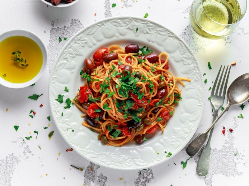 Spaghetti alla Puttanesca auf einem weißen, gemusterten Teller. Garniert sind sie mit Petersilie und daneben liegen Gabel und Löffel sowie Olivenöl und ein Glas Weißwein.