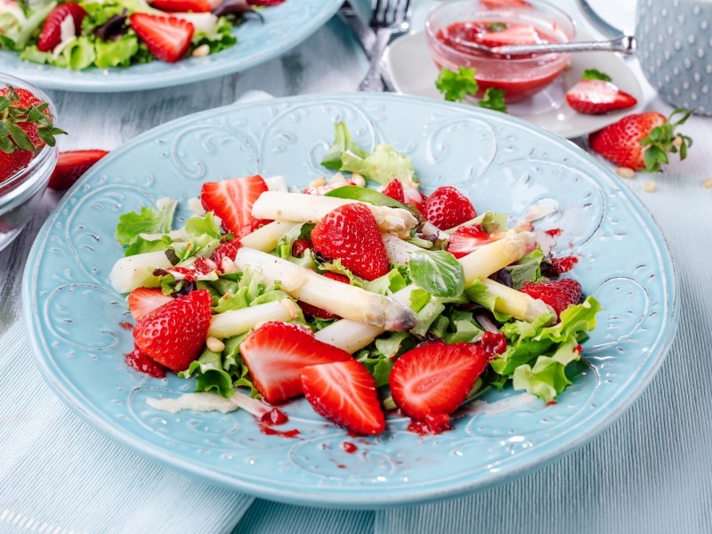 Spargel-Erdbeer-Salat auf einem hellblauen, gemusterten Teller. Im Hintergrund stehen mehr Teller mit Salat.