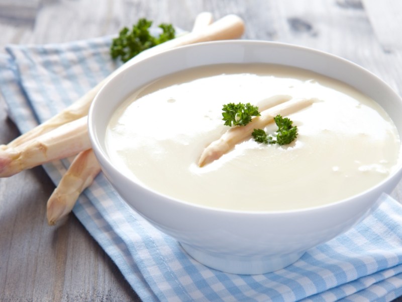 Eine Schüssel Spargelcremesuppe mit Petersilie. Frische Spargelstangen liegen daneben.