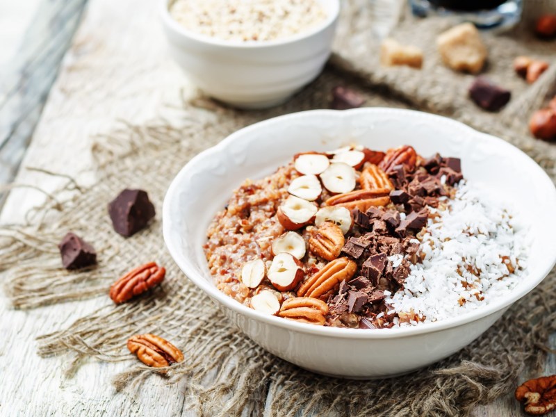 Süße Quinoa-Bowl in weißer Schüssel, garniert mit Haselnüssen, Pekannüssen, Kokosraspeln und Schokolade.