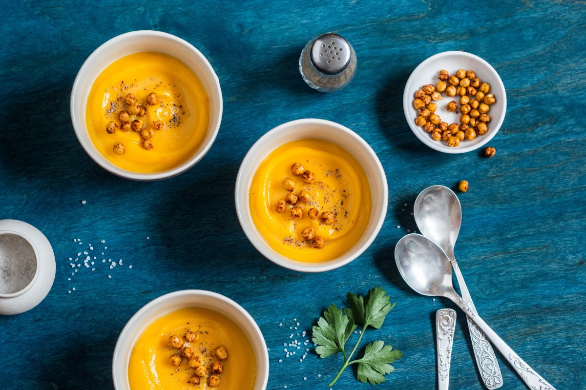 Drei Schalen mit orangefarbener Süßkartoffelsuppe mit gerösteten Kichererbsen und Löffel daneben auf blauem Untergrund.