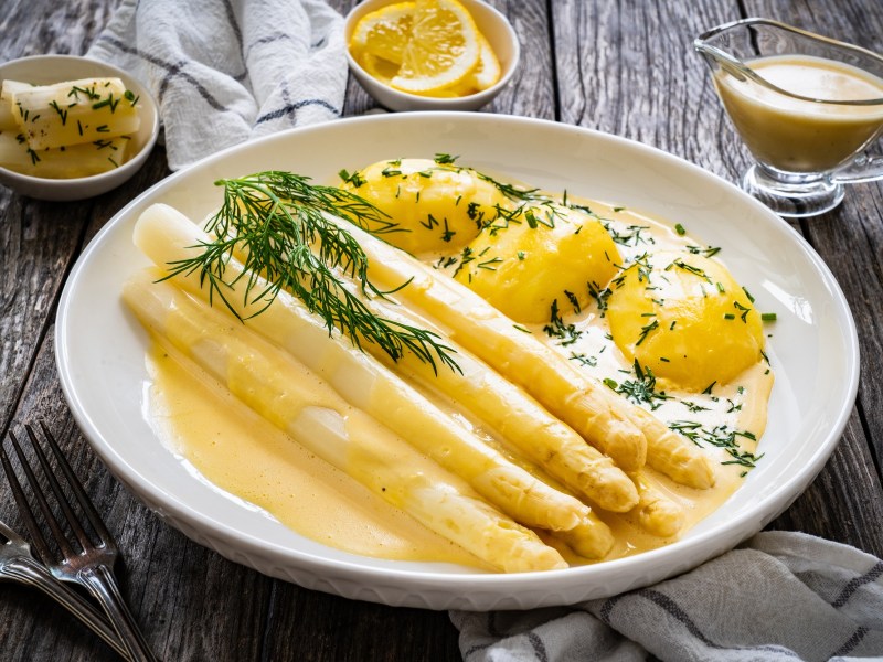 ein Teller weißer Spargel mit veganer Sauce hollandaise und Kartoffeln und Petersilie, daneben je eine Schale mit Butter, mit Zitronen und Soße.