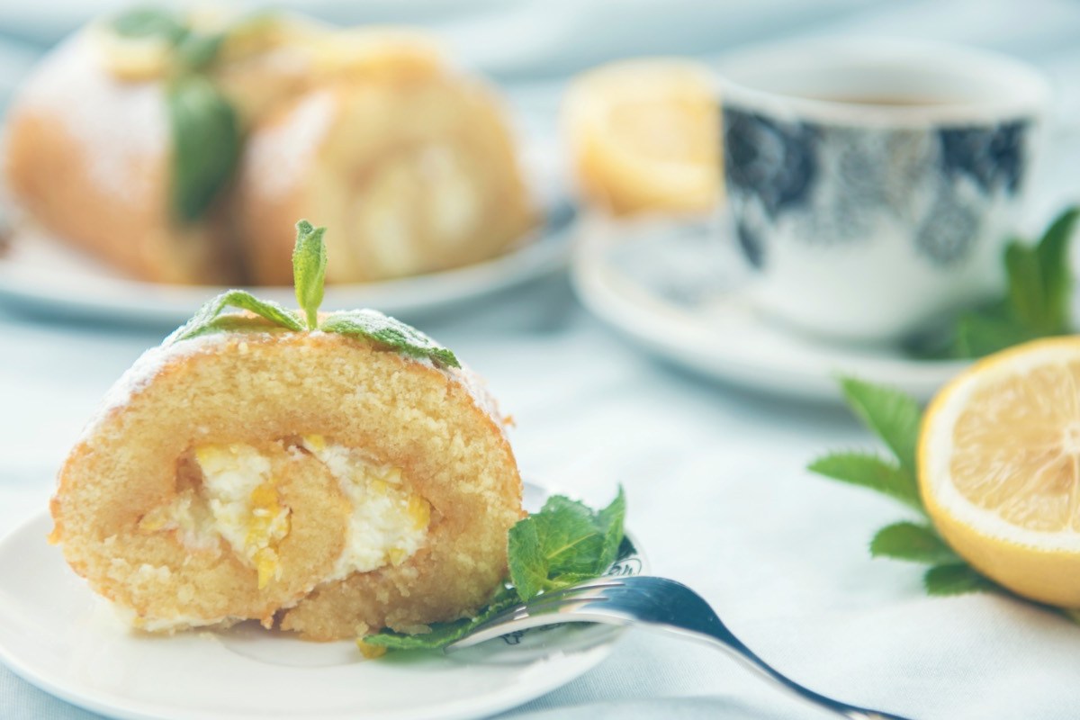 Ein Stück Zitronen-Biskuitrolle, mit Minze dekoriert, auf einem Teller. Im Hintergrund sind eine Tasse Kaffee und der restliche Kuchen zu sehen.