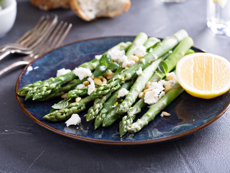 Gegrillter Spargel mit Feta und Zitrone sowie Pinienkernen auf einem Teller, daneben Besteck.