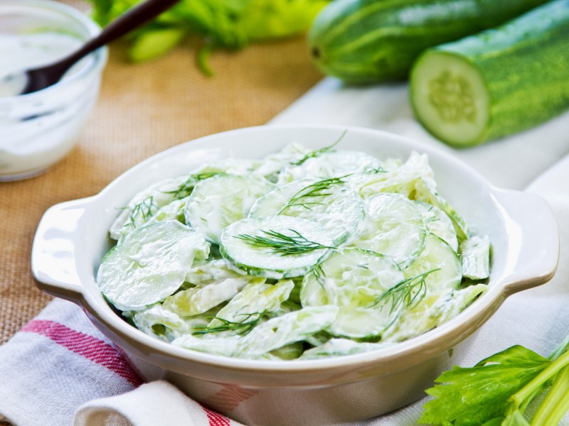 Eine Schüssel Gurkensalat mit Sahne, dahinter unscharf eine Schüssel mit der Salatsoße und Gurken.