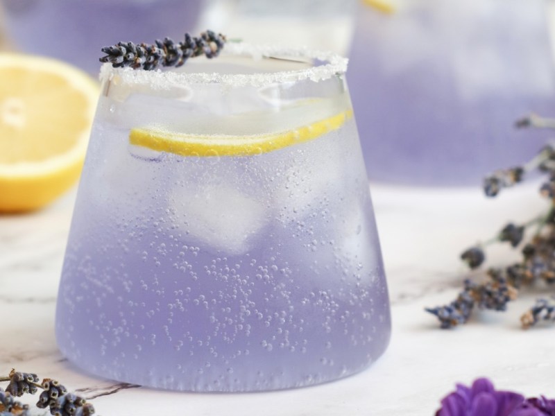 Ein Glas Holunder-Lavendel-Spritz. Drumherum liegen Lavendelblüten und eine halbe Zitrone.