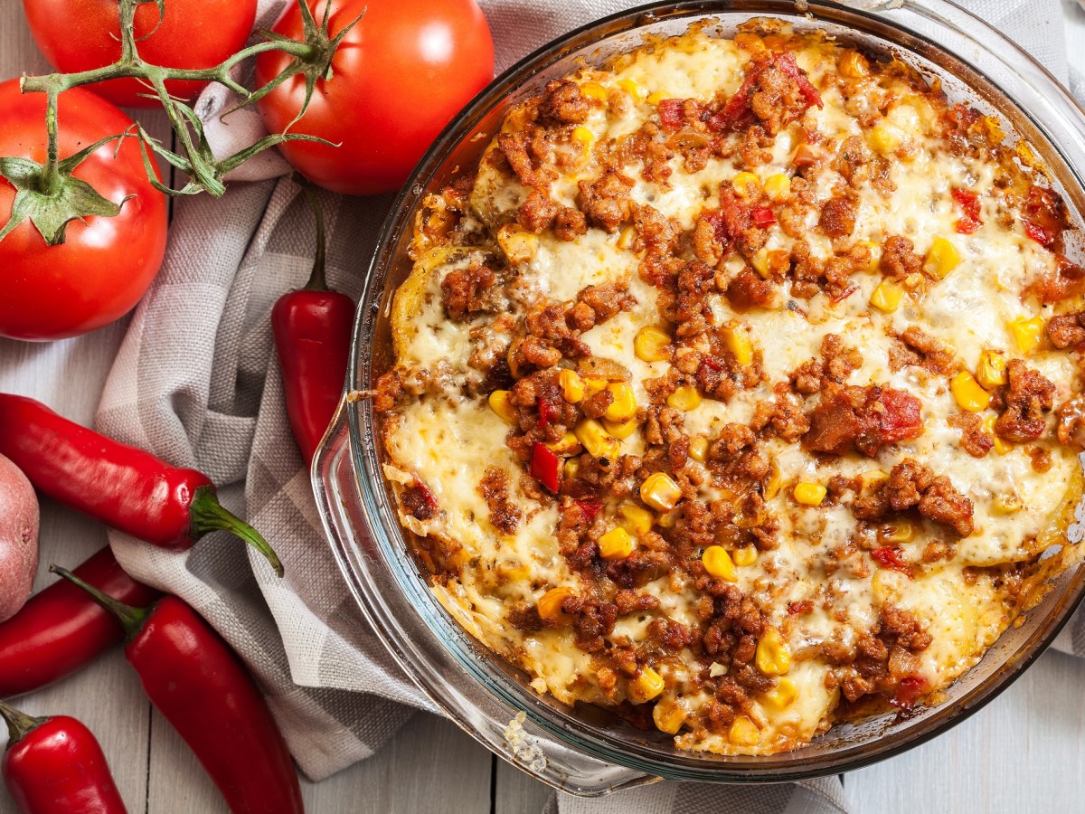Kartoffel-Hack-Auflauf nach mexikanischer Art in einer Auflaufform, daneben Tomaten, Chilis und Kartoffeln.