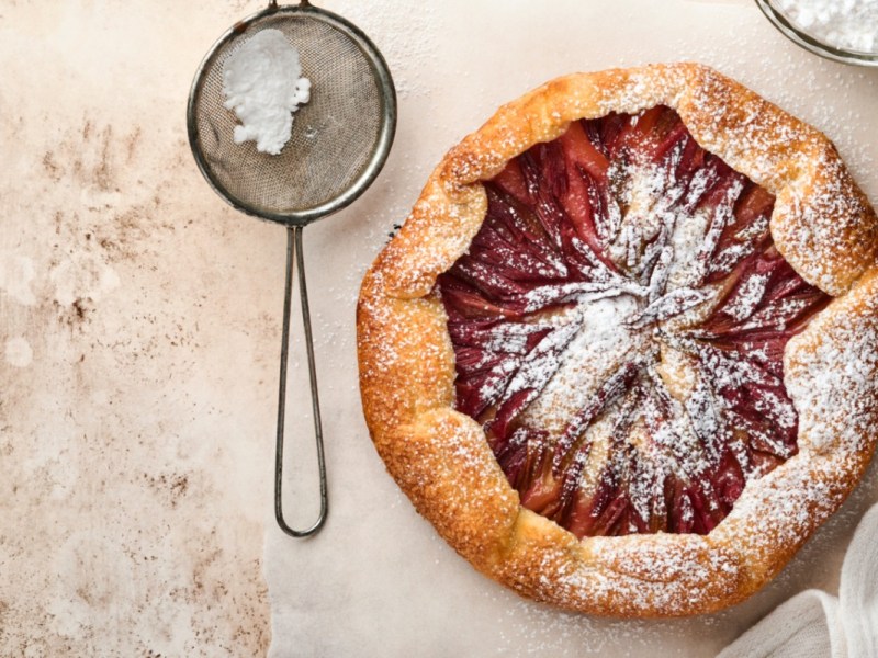 Eine Rhabarber-Galette mit Puderzucker in der Draufsicht. Daneben liegt ein Sieb mit Puderzucker.