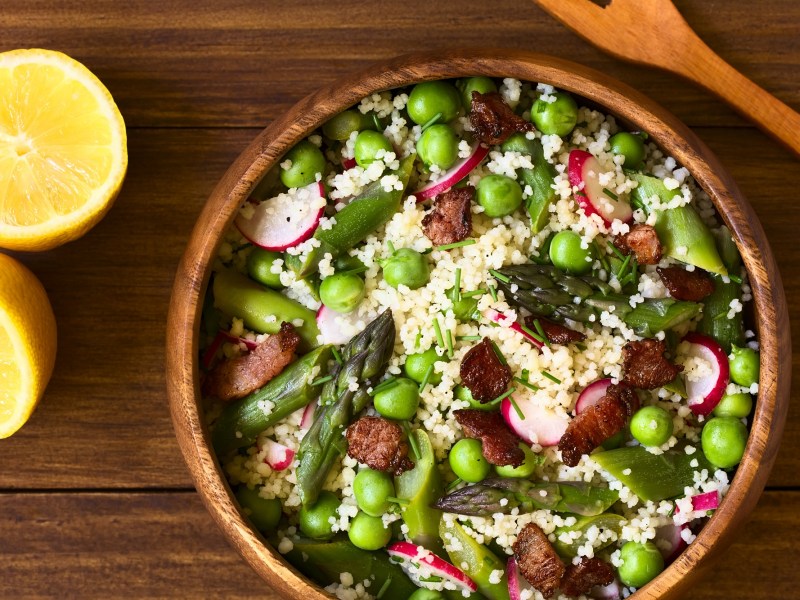 Eine Holzschüssel (Öffnet in einem neuen Browser Tab) Spargel-Couscous-Salat mit Erbsen und Radieschen in der Draufsicht, daneben eine halbierte Zitrone und ein Holzlöffel.