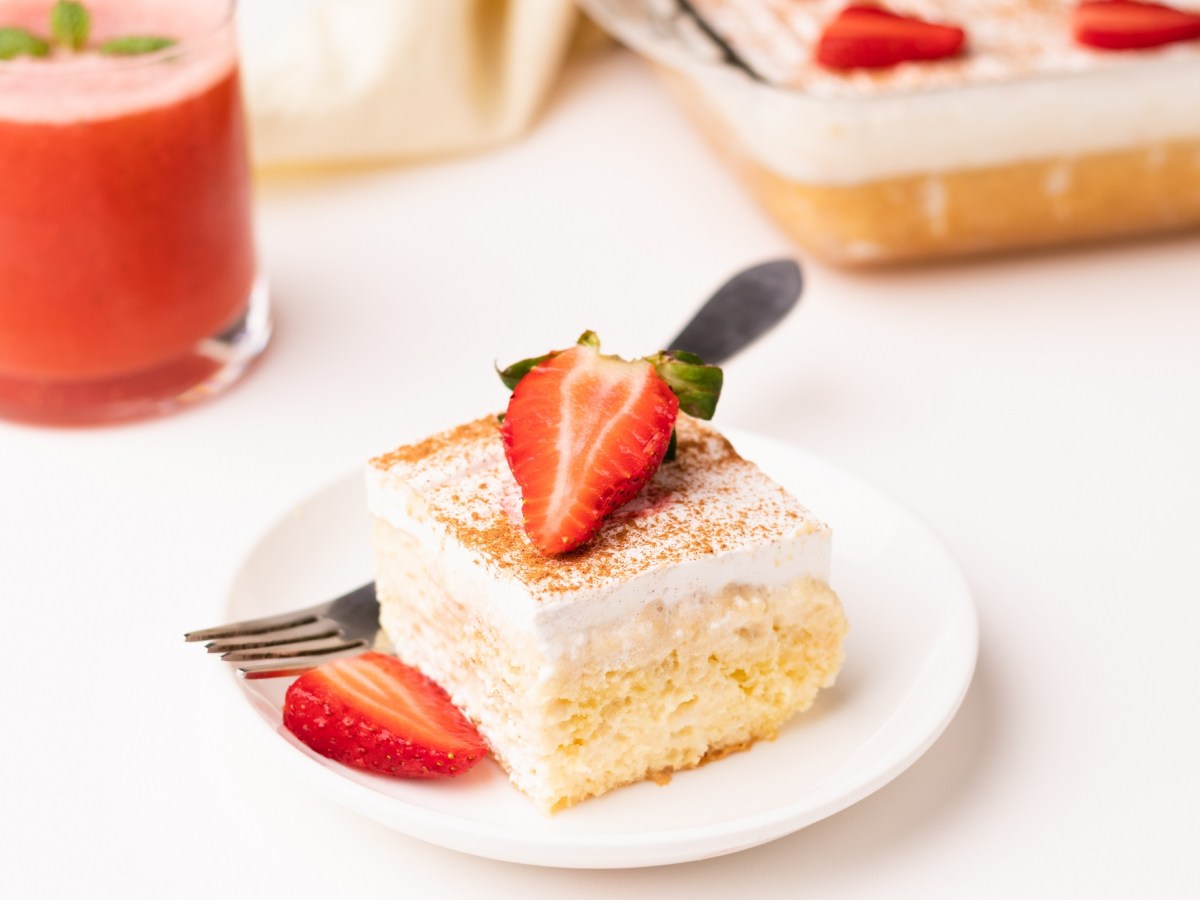 ein Stück Tres Leches Kuchen mit Erdbeeren auf einem weißen Teller. Dahinter steht unscharf der Rest des Kuchens