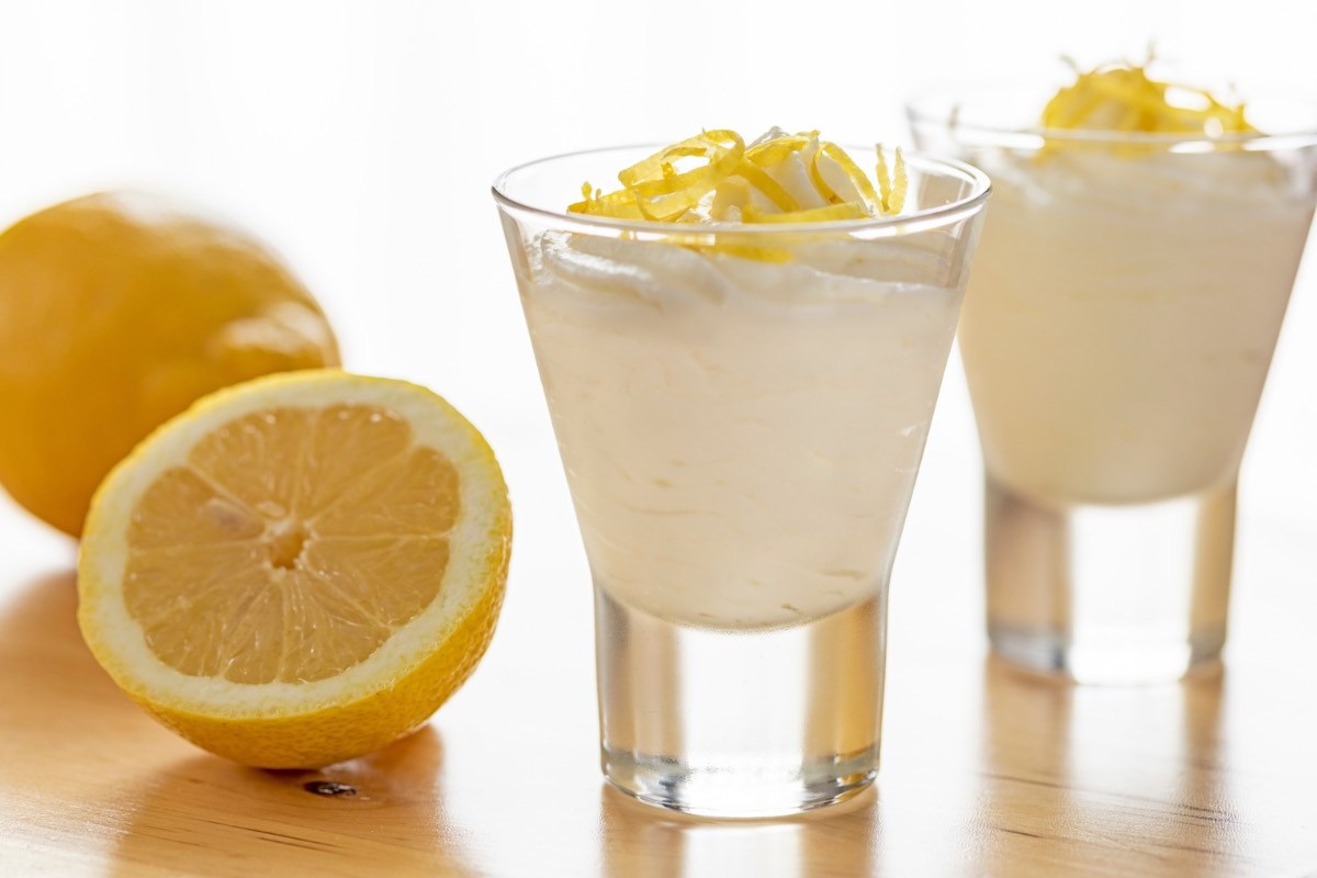 2 Gläser mit Zitronenmousse. Daneben liegt eine halbierte, frische Zitrone.