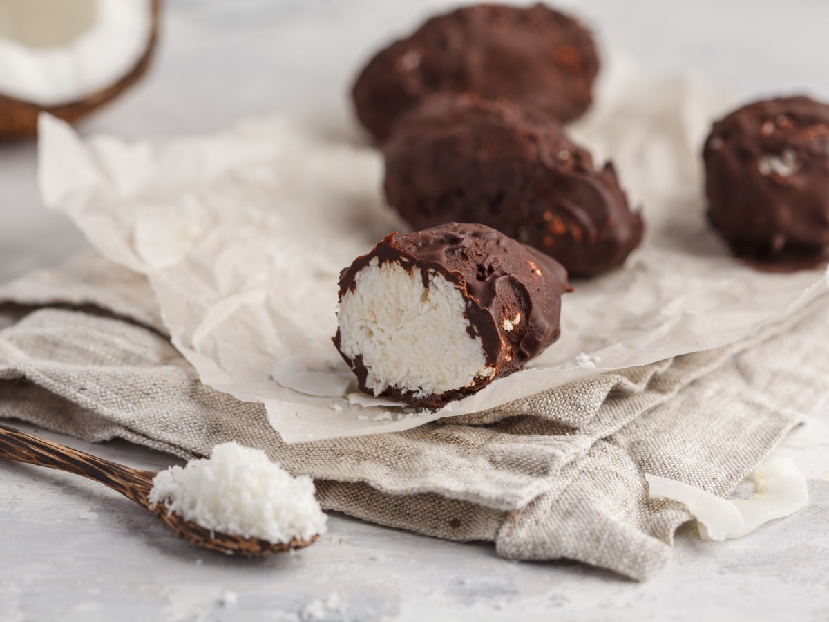 vegane Bounty mit Schokoglasur auf einem Stück Papier, daneben eine halbe Kokosnuss und ein Löffel Kokosraspeln.