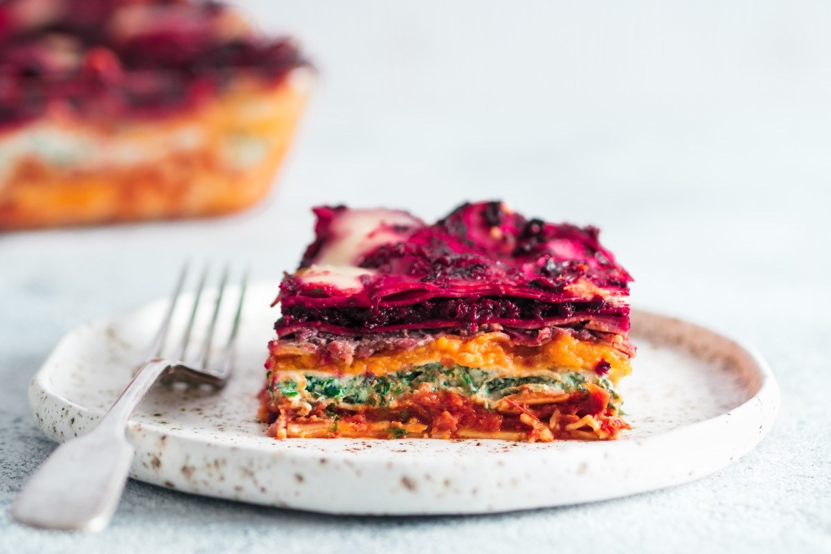 Ein Stück vegane Regenbogen-Lasagne auf einem Teller. Der Rest ist im Hintergrund zu sehen.