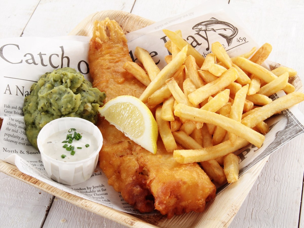 Fish and Chips in einer Schale, auf Zeitungspapier angerichtet. Dabei Erbsenpüree, Remoulade und Zitrone.