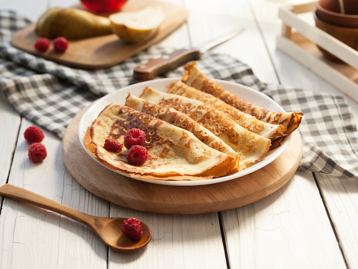 Einige Kefir-Pfannkuchen auf einem Teller, garniert mit Himbeeren, daneben einige Zutaten.