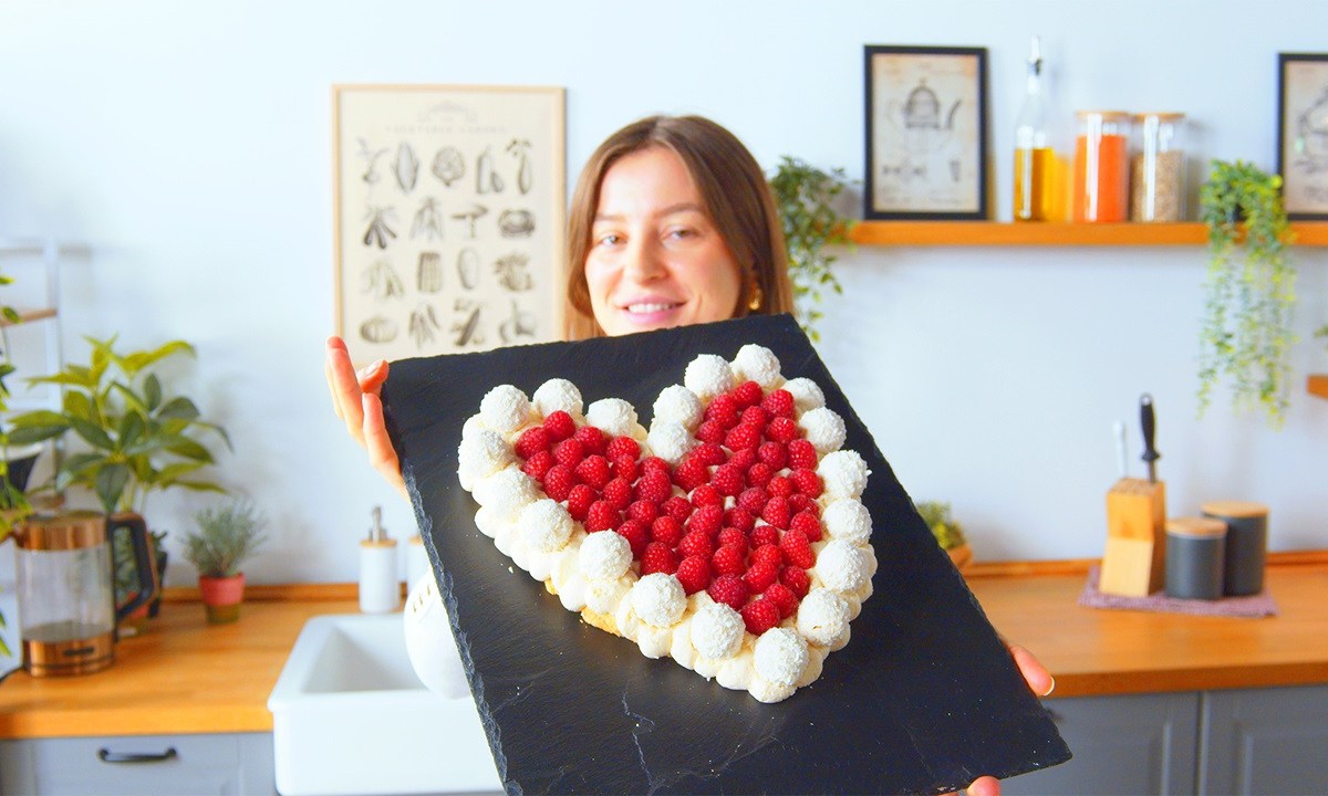 Unsere Köchin hält den Raffaello-Herzkuchen auf einer Schieferplatte in die Kamera. Der Raffaello-Herzkuchen besteht aus weichen Löffelbiskuits, ist am Rand von Raffaellos umgeben und mit Himbeeren dekoriert. Im Hintergrund sieht man eine Kochzeile mit, von links nach rechts, einem Wasserkocher, Spüle, einem Messerblock sowie Aufbewahrungsdosen.