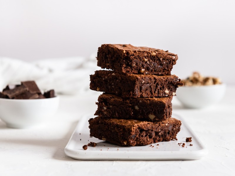 Ein Stapel zuckerfreie Apfel-Brownies auf einem weißen, eckigen Teller. Im Hintergrund stehen eine Schüssel Schokolade und eine mit Walnüssen.