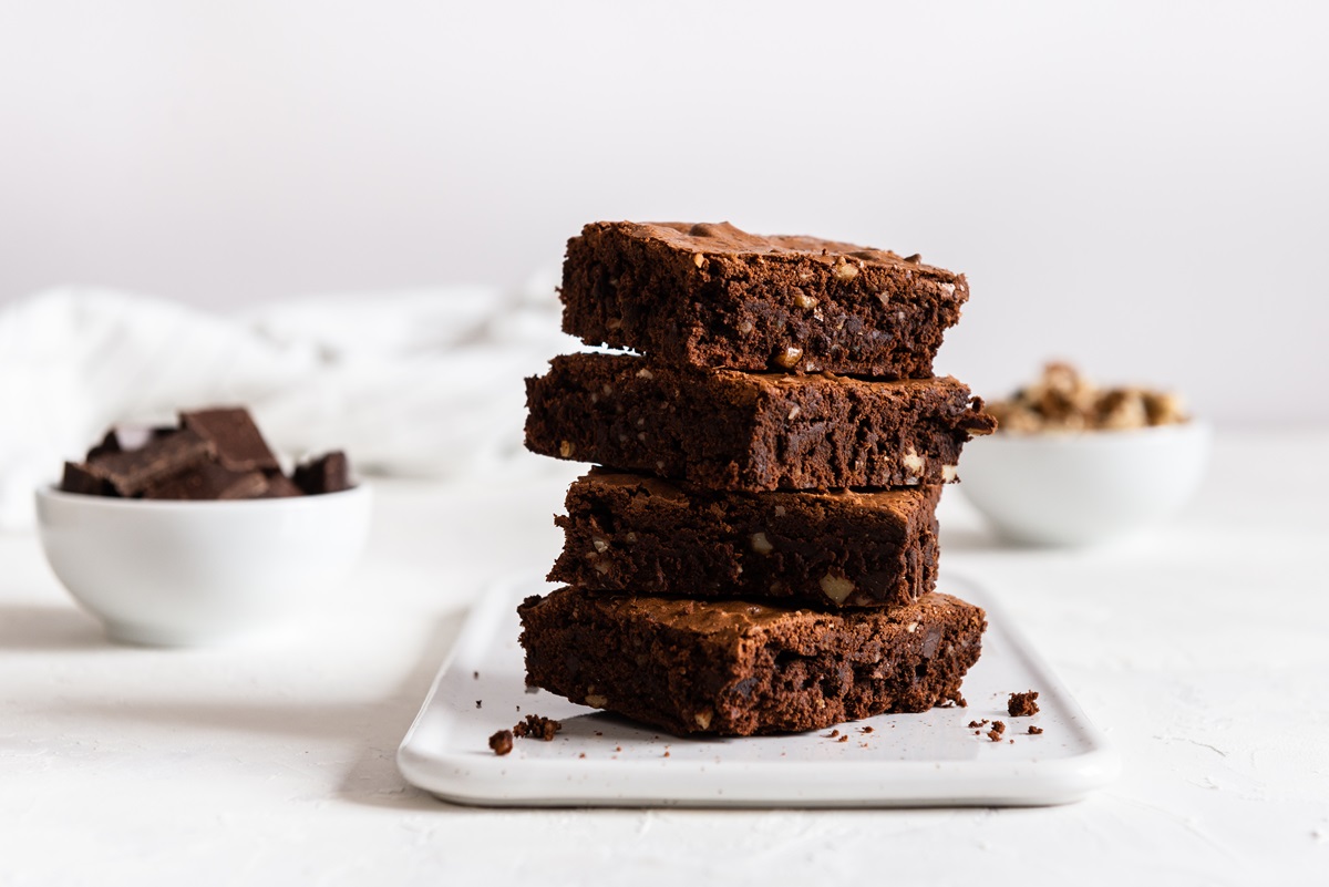 Ein Stapel zuckerfreie Apfel-Brownies auf einem weißen, eckigen Teller. Im Hintergrund stehen eine Schüssel Schokolade und eine mit Walnüssen.