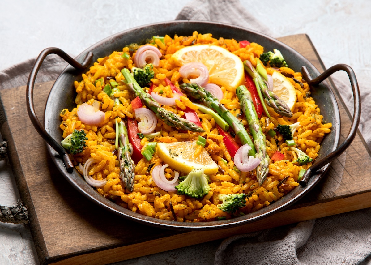 Mediterrane Reispfanne mit Spargel, Brokkoli, Paprika und Zwiebel in einer gusseisernen Pfanne, die auf einem Holzbrett steht. Nahaufnahme.