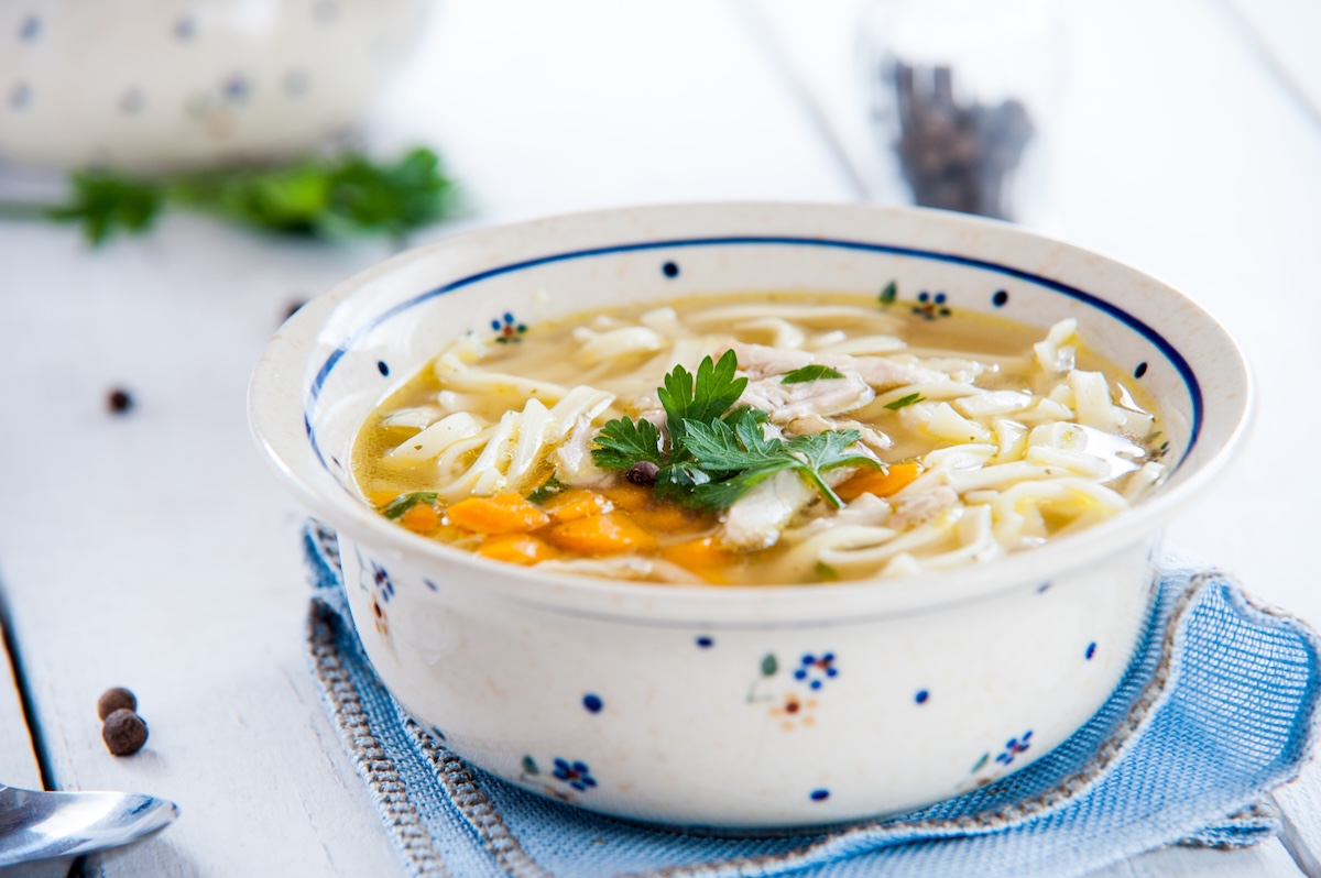 Eine Schüssel Nudelsuppe mit Gemüse auf einer Stoffserviette, daneben ein Löffel.