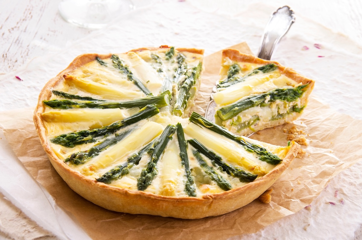 Eine runde, angeschnittene vegane Spargelquiche, aus der ein Stück herausgenommen wird, auf einem Stück Backpapier.