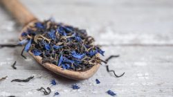 Earl Grey selbst machen: Ein Löffel Earl Grey mit Kornblumenblüten liegt auf einem hellen Holztisch.
