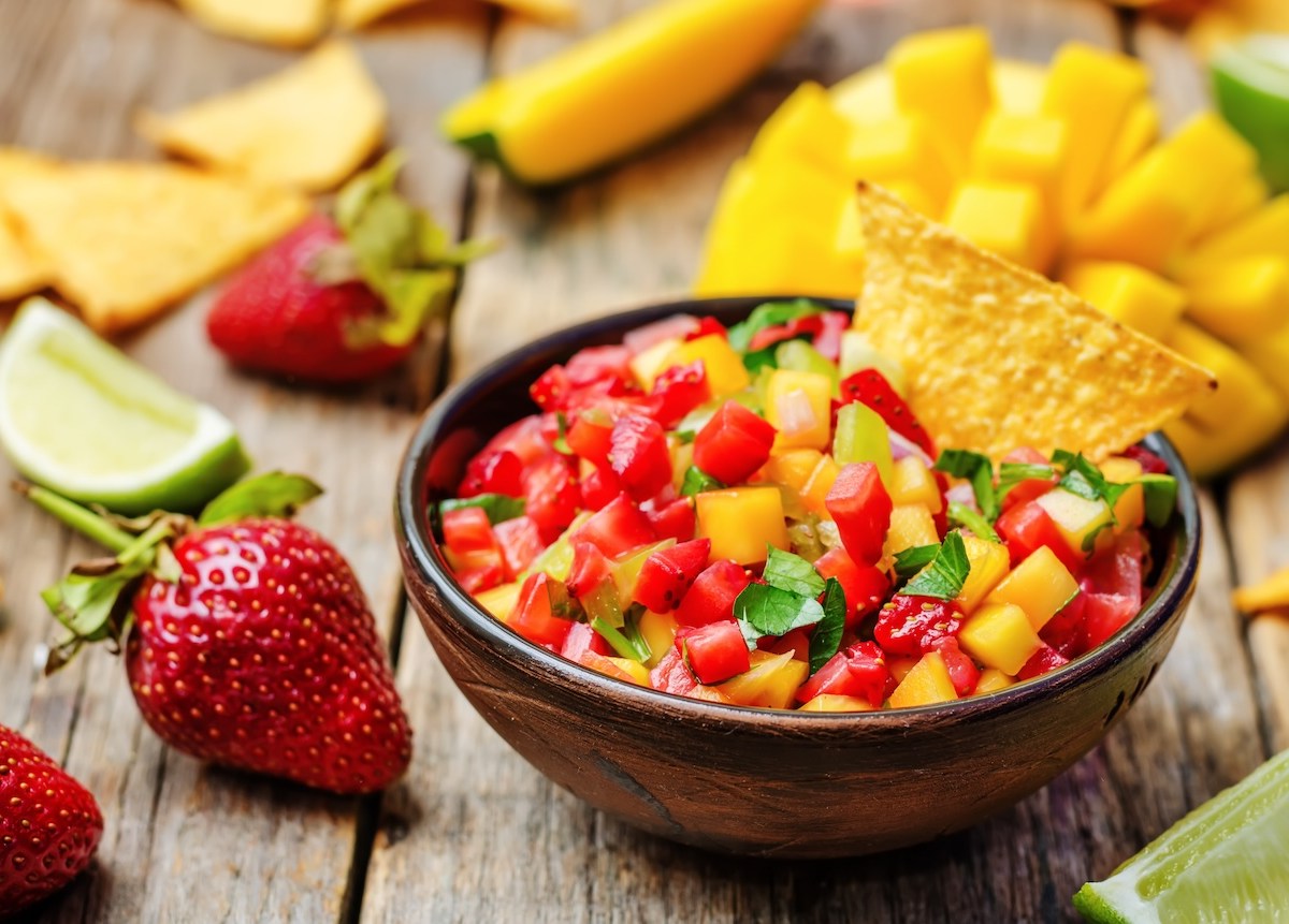 Eine Schale Erdbeer-Mango-Salsa mit einem Nacho, drumherum Erdbeeren, Mango und Limettenspalten.