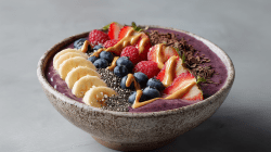 Eine Beeren-Quark-Bowl auf grauem Untergrund. Die Bowl besteht aus lila farbenen Joghurt und ist mit frischen Bananen, Erdbeeren sowie gefrorenen Heidelbeeren und Himbeeren angerichtet. Etwas Nussmus, Chiasamen und Schokoraspeln sind ebenfalls darübergestreut.