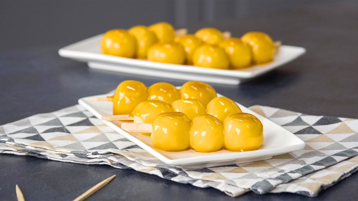 Drei Dango-Spieße liegen auf einem weißen Teller. Im Hintergrund sieht man weitere Dango-Spieße. Sie sind mit einer goldfarbenen Glasur übergossen.