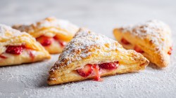 Ein paar Erdbeer-Blätterteigtaschen mit Vanille-Creme, die mit Puderzucker bestäubt sind. Auf der Arbeitsfläche liegt auch Puderzucker verstreut.