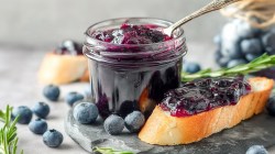 Ein Einmachglas mit herzhafter Blaubeer-Zwiebel-Marmelade, in der ein Löffel steckt. Neben dem Glas liegt eine Scheibe Baguette, die mit der Marmelade bestrichen ist. Ringsum liegen einzelne Blaubeeren herum.