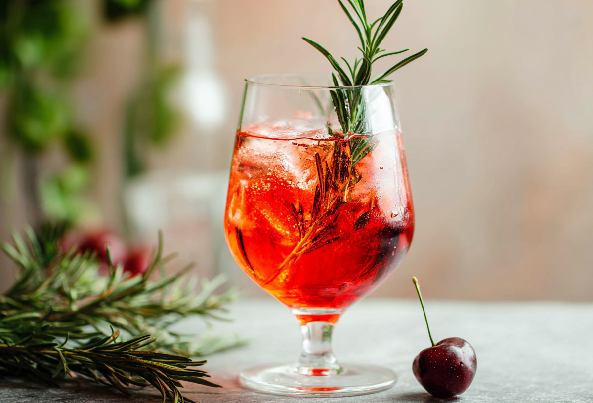 Ein Glas Savoia Spritz, garniert mit Rosmarin und einer Kirsche.