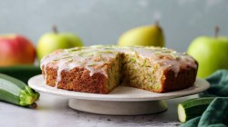 Ein runder Apfel-Zucchini-Kuchen, der auf einer Kuchen-Etagere liegt. Ringsum liegen frische Äpfel und Zucchini verteilt.