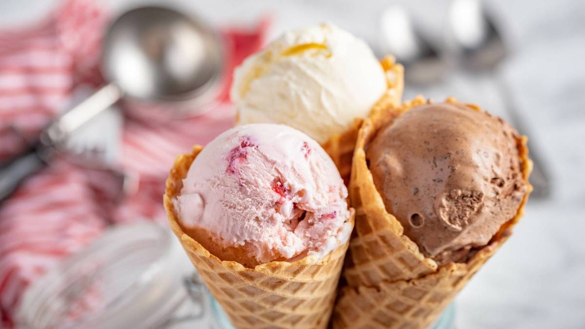 Drei Eiswaffeln mit jeweils einer Kugel Eis darin. Dabei handelt es sich um Erdbeer-, Vanille und Schokoladeneis, das mit der KLAMER 2-in-1 Eismaschine hergestellt wurde. Im Hintergrund kann man einen Eisportionierer sowie 2 Löffel sehen. Der Portionierer liegt auf einem rot-weißen Küchenhandtuch.