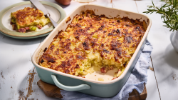 Eine Auflaufform mit einem Döppekuchen steht auf einem Brett auf einem blauen Tuch. Links im Hintergrund ist ein Teller mit einem Stück des Auflaufs. Rechts im Bild sieht man frischen Rosmarin.